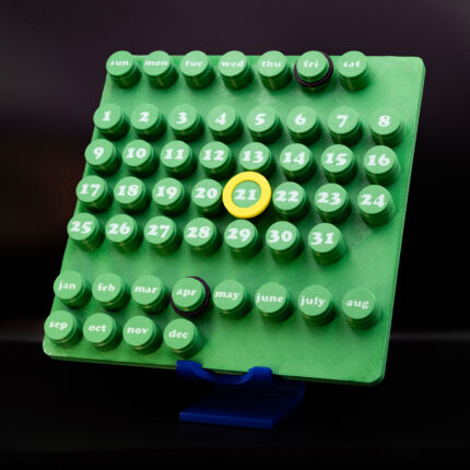 3D printed modular desk calendar with green layout and movable date markers placed on a modern wooden console with lamp and decor