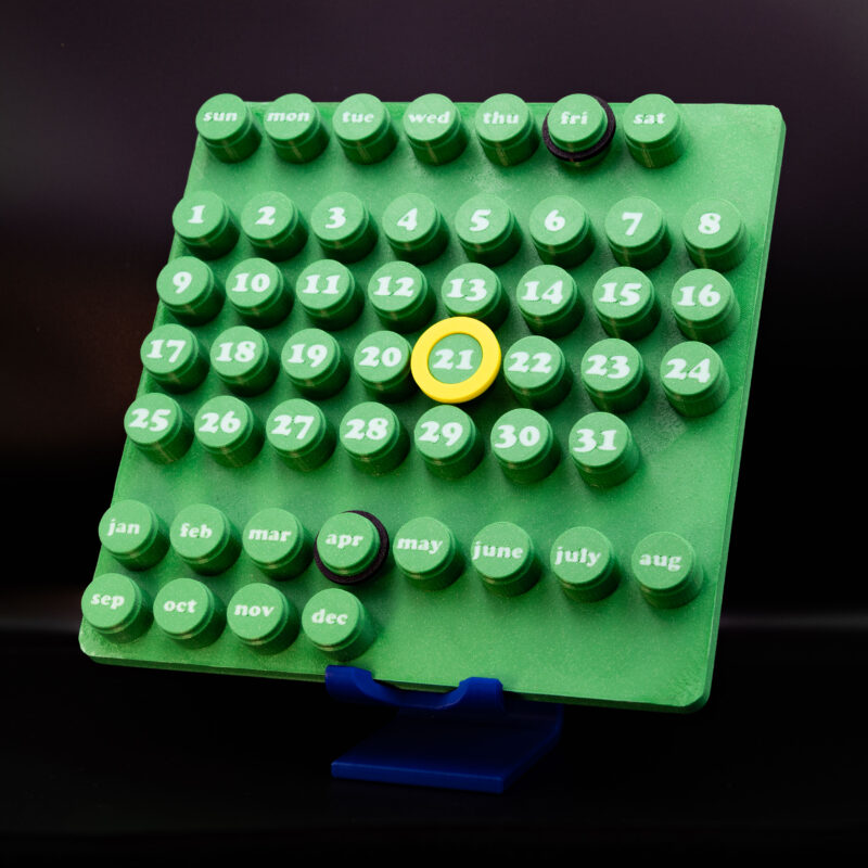 3D printed modular desk calendar with green layout and movable date markers placed on a modern wooden console with lamp and decor